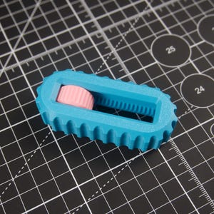 May include: A blue and pink 3D-printed rotary cutter. The cutter has a ridged blue body with a pink, cylindrical roller. The cutter is on a black cutting mat with white grid lines and circular markings.