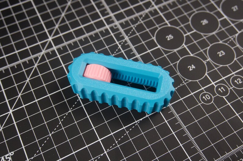 May include: A blue and pink 3D-printed rotary cutter. The cutter has a ridged blue body with a pink, cylindrical roller. The cutter is on a black cutting mat with white grid lines and circular markings.