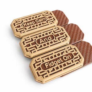 May include: Three wooden fidget toys with the phrases "Keep Calm", "And", and "Fidget On" engraved on them. The fidget toys have a light brown frame with a darker brown center. The background is white.