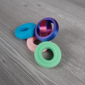 May include: Four colorful finger exercisers in blue, pink, green, and a gradient of blue to purple. The exercisers have a ridged texture and a circular shape. The background is a gray wood grain.