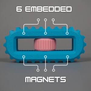 May include: A turquoise blue, gear-shaped object with a rectangular center, housing a pink cylinder. The text "6 EMBEDDED MAGNETS" is displayed above and below the object, with lines pointing to the embedded magnets.