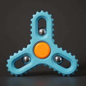 May include: A blue fidget spinner with a gear-like design. It has three arms, each with a silver ball bearing. The center features an orange circle. The background is a solid gray.