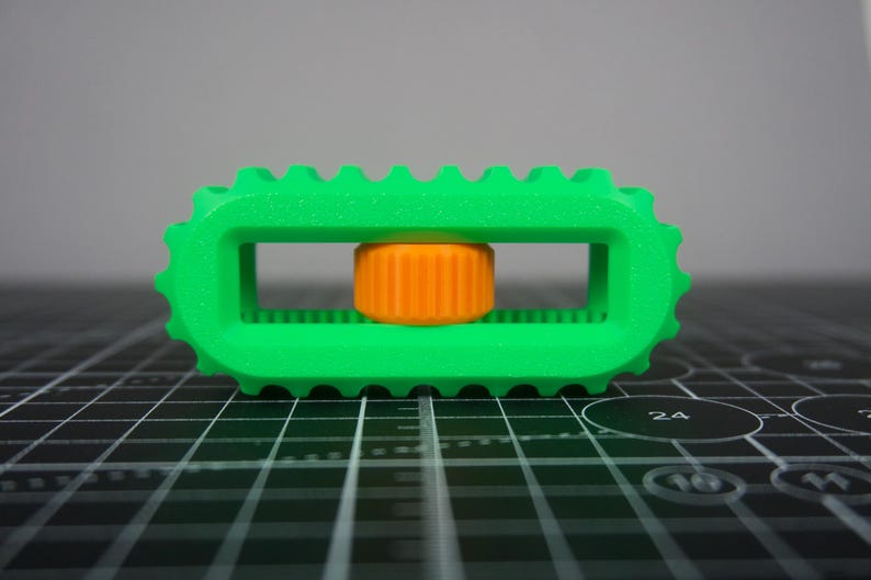 May include: A bright green, rectangular 3D-printed object with a ridged edge and a central orange cylinder. The object has a geometric design and appears to be a tool or component.