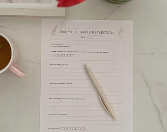 May include: A Daily Check-In & Reflection worksheet with prompts for self-reflection. A pen with a dotted pattern lies on the page. A pink mug and a red tulip are in the background. The worksheet is on a white surface.