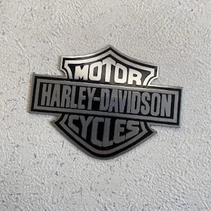 May include: A silver and black Harley-Davidson Cycles emblem. The emblem features the words "MOTOR HARLEY-DAVIDSON CYCLES" in a rectangular and shield-shaped design. The emblem is set against a textured, off-white background.