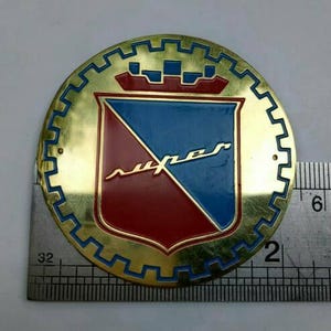 Può includere: Un emblema rotondo color oro con un design a scudo blu e rosso. Lo scudo presenta la parola "super" in corsivo. L'emblema misura circa 5 cm di diametro e ha un bordo blu e oro.