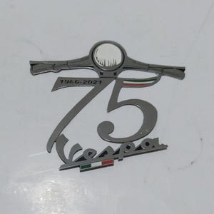 Puede incluir: Un emblema plateado del 75 aniversario de Vespa. El emblema presenta el número 75, el logotipo de Vespa y los años 1946-2021. El emblema también incluye un diseño de manillar y los colores de la bandera italiana.