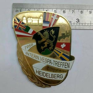 Peut inclure: Une épingle dorée vintage en forme de bouclier. Elle présente des drapeaux, un blason de lion et le texte "1959 INTERN.VESPA-TREFFEN HEIDELBERG." L'épingle mesure environ 7.6 cm de long, comme mesuré par la règle.
