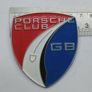 May include: A vintage Porsche Club emblem with a shield shape. The emblem features red, white, and blue sections, with the words "PORSCHE CLUB" in white. The letters "GB" are in silver. The emblem is approximately 3 inches wide.