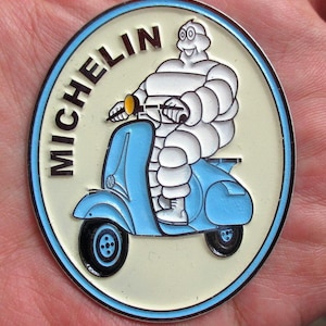 May include: A round, cream-colored emblem with a blue border featuring the Michelin Man riding a light blue scooter. The word "MICHELIN" is printed in black around the top of the emblem. The Michelin Man is white with black details.