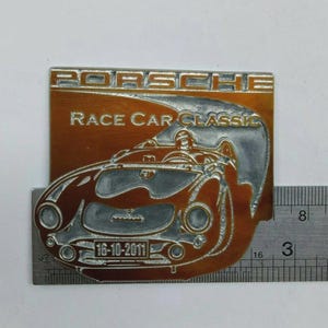 May include: A rectangular metal emblem with the word "PORSCHE" at the top and "RACE CAR CLASSIC" below. The emblem features a detailed illustration of a vintage race car in silver against a copper-colored background. The date "16-10-2011" is also visible.