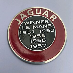 May include: A circular metal emblem featuring a red outer ring with the word JAGUAR in silver lettering. The black centre displays the text WINNER LE MANS followed by the years 1951, 1953, 1955, 1956, and 1957 in raised silver-coloured characters.
