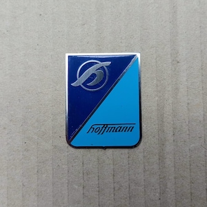 Peut inclure: Un badge rectangulaire Hoffmann avec une bordure argentée. Le badge présente un fond bleu avec une section diagonale bleu clair. Une lettre "h" argentée stylisée est en haut, et le nom "hoffmann" est imprimé en noir.
