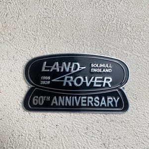 May include: Black and silver Land Rover 60th Anniversary emblem. The top section reads "LAND ROVER" with "SOLIHULL ENGLAND" below. The bottom section reads "60TH ANNIVERSARY" with the years "1966 2026" on the top.
