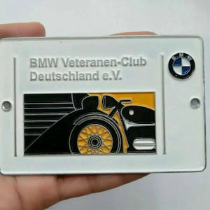 Puede incluir: Un letrero rectangular de metal blanco con el texto "BMW Veteranen-Club Deutschland e.V." en gris. El letrero incluye un logotipo de BMW y un gráfico de una motocicleta negra y amarilla. Se ven dos pequeños agujeros.