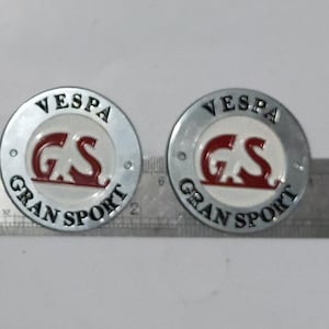 May include: Two Vespa Gran Sport emblems. Each emblem is a silver-colored circle with the word "VESPA" at the top. The center features "GS" in red, with "GRAN SPORT" around the bottom. The emblems are on a white surface.