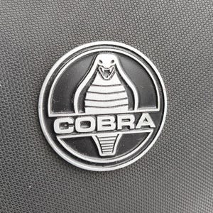 May include: A circular, silver-rimmed emblem featuring a black cobra design with the word "COBRA" in white block letters. The cobra is centered, with its head raised and fangs bared. The emblem is set against a textured, dark gray background.