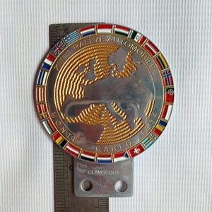 Puede incluir: Una insignia metálica plateada y dorada con el texto "XIII RALLYE AUTOMOBILE MONTE CARLO 1934". La insignia presenta un mapa del mundo de color dorado y está rodeada por un anillo de banderas internacionales. Un soporte metálico está adjunto en la parte inferior.
