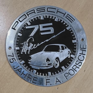Op de afbeelding: Een cirkelvormig, zilverkleurig metalen embleem met het woord "PORSCHE" rond de buitenrand. Het midden toont het nummer "75", een auto-illustratie en de tekst "75 Jahre F. A. Porsche".