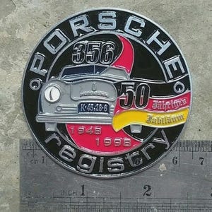 May include: A circular metal emblem with the word "PORSCHE" in silver lettering. The emblem features a vintage car, the number "356", and the text "registry". It also includes the years "1948" and "1998".