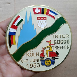 Puede incluir: Un pin metálico redondo de color crema con borde dorado. El pin presenta una cordillera azul, un río verde y banderas. El texto incluye "INTER GOGGO TREFFEN" y "KÖLN 6.-7. JUNI 1953" con un gráfico de scooter.