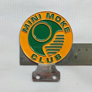 May include: A round, orange and green Mini Moke Club emblem with the text "MINI MOKE CLUB" in green. The emblem has a silver metal mounting bracket attached to the bottom. A ruler shows the emblem is approximately 7.6 cm wide.