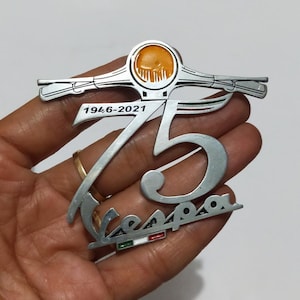 Può includere: Un emblema Vespa argentato che commemora 75 anni, dal 1946-2021. L'emblema presenta il logo Vespa, un design stilizzato del manubrio dello scooter e il numero 75. L'emblema è tenuto in una mano.