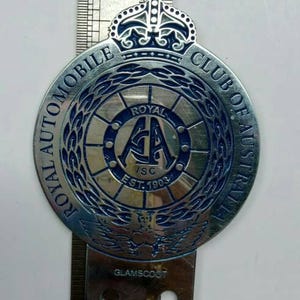 May include: A silver-colored Royal Automobile Club of Australia badge with blue lettering and accents. The badge has a crown at the top and the text "ROYAL AUTOMOBILE CLUB OF AUSTRALIA" around the edge. The center features the letters "RAA" and "ISC EST. 1903".
