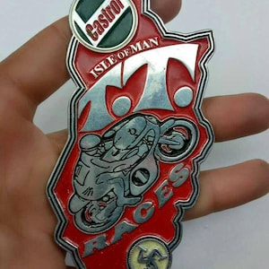 May include: A red, silver, and green Isle of Man TT Races emblem. The emblem features the Castrol logo, the text "Isle of Man", and a motorcycle racing graphic. The emblem is shaped like the Isle of Man.