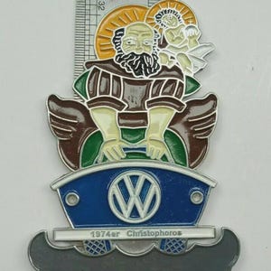 May include: A vintage metal pin featuring a religious figure with a halo, seated above a blue VW logo. The pin includes the text "1974er Christophoros" and is approximately 2 inches in height.