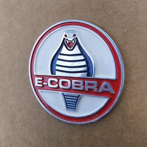 May include: A round, metal emblem with a red border and the text "E-COBRA". The emblem features a white background with a blue and white cobra design in the center. The cobra has a red tongue.