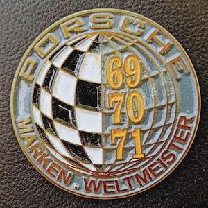 May include: A round, vintage Porsche emblem with a gold border. The emblem features the word "PORSCHE" at the top, and "MARKEN WELTMEISTER" around the bottom. The center shows a globe with a black and white checkered pattern and the numbers 69, 70, and 71.