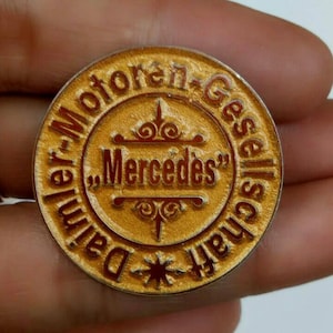 Può includere: Una spilla rotonda color oro con le parole "Daimler-Motoren-Gesellschaft" che circondano il bordo. La parola "Mercedes" è al centro, in lettere rosse. Un disegno a stella è in basso.