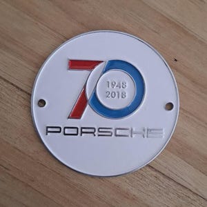 May include: A white, circular Porsche emblem with a red and blue "70" design, the years "1948" and "2018" inside a blue circle, and the brand name "PORSCHE" in silver lettering. Two small holes are visible.