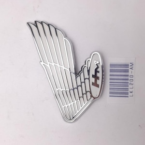 May include: A silver-colored wing-shaped emblem with white detailing. The emblem has a stylized wing design with a series of parallel lines. The emblem also features a small, oval-shaped section with the letters "HM" in a brown color. A barcode sticker is visible.