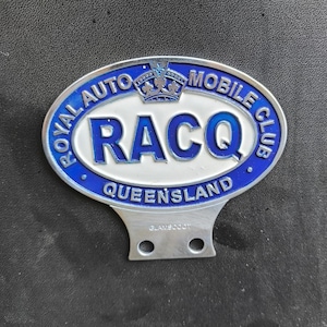 May include: An oval RACQ Royal Auto Mobile Club emblem in blue and white. The emblem has the text "ROYAL AUTO MOBILE CLUB" and "QUEENSLAND" in blue, with "RACQ" in bold blue letters. A silver metal base is attached.