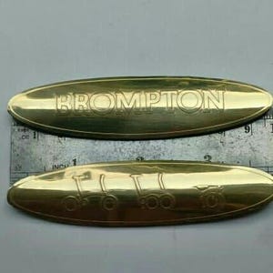 May include: Two gold-colored, oval-shaped metal plates. The top plate has the word "BROMPTON" embossed on it. The bottom plate features an embossed design of a bicycle. A ruler with inch and cm measurements is in the background.