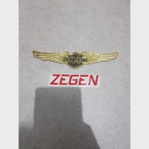 May include: A gold-colored Harley-Davidson Cycles emblem with wings, and the word "ZEGEN" in red letters on a white background. The emblem is a motorcycle accessory.