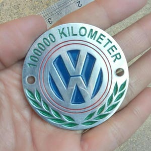 May include: A silver-colored, circular emblem with the text "100000 KILOMETER" in green. The center features the Volkswagen logo in blue. A green laurel wreath design is at the bottom.