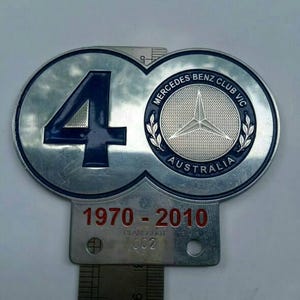May include: A silver-colored Mercedes-Benz Club VIC Australia emblem. The emblem features the number 40 in blue, the Mercedes-Benz logo, and the text "1970-2010". The emblem is attached to a rectangular metal plate.