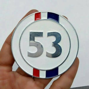 Può includere: Un emblema rotondo in metallo bianco con il numero "53" in argento. L'emblema ha un bordo argentato e strisce rosse e blu nella parte superiore. L'emblema è tenuto in una mano.