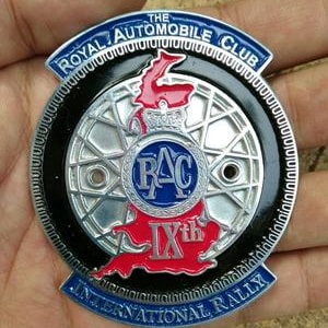 May include: A circular metal emblem with the text "THE ROYAL AUTOMOBILE CLUB" and "INTERNATIONAL RALLY". The emblem features a wheel design, a red map outline, and the letters "RAC" in a blue circle. The text "IXth" is also visible.