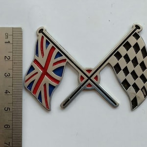 May include: A silver-colored metal emblem featuring two crossed flags. One flag is the Union Jack, with red and blue accents. The other is a black and white checkered flag. The emblem is approximately 2 inches wide.