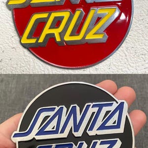 May include: Two circular metal emblems featuring the Santa Cruz logo in raised lettering. The top emblem has a red background with yellow text, while the bottom emblem has a black background with blue text. Both feature a silver metallic border.