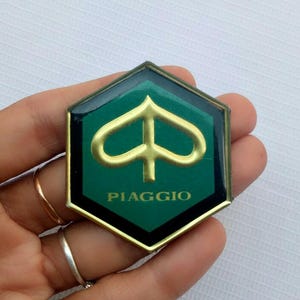 Può includere: Un emblema Piaggio verde e oro con un design a forma di picche. L'emblema è esagonale con un bordo nero e la scritta "PIAGGIO" in bianco.