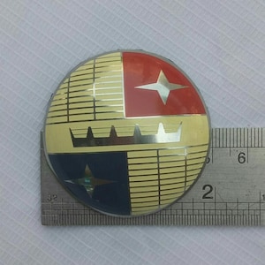 Può includere: Un emblema rotondo vintage con un design segmentato. Presenta quadranti rossi e blu, ciascuno con una stella bianca. Gli altri due quadranti sono gialli con dettagli a corona argentata e linee orizzontali. Diametro di circa 5 cm.