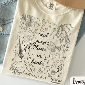 Puede incluir: Una camiseta de color marfil con un dise&ntilde;o gr&aacute;fico negro que presenta criaturas m&iacute;ticas y el texto "real magic lives in books". La camiseta se combina con vaqueros azul claro, un collar dorado y un sombrero beige.
