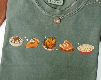 Può includere: T-shirt verde muschio con una grafica di cibo del Ringraziamento: purè di patate, torta, tacchino arrosto, panini e mac and cheese. Una collana d'argento con un ciondolo è drappeggiata sulla maglietta. La maglietta è in Comfort Colors.