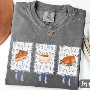 May include: A gray Comfort Colors t-shirt featuring three square patches with Thanksgiving-themed illustrations. The patches include a slice of pie, gravy, and a roasted turkey. Each patch has a blue floral pattern and a blue bow.