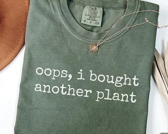 Peut inclure: Un t-shirt vert mousse avec le texte "oops, i bought another plant" en blanc. Un collier doré avec un pendentif repose sur le t-shirt. Un chapeau marron et des tiges de plantes séchées sont également visibles.